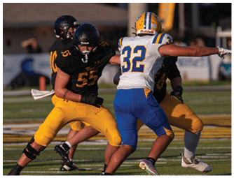 Two people in football uniforms are engaged in a play on a grassy field with one person in black and yellow and the other in white, blue, and yellow. Descripci n generada con IA