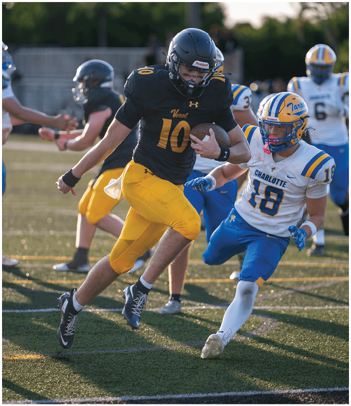 A person in a black and yellow football uniform is running with the ball while being pursued by two people in white and blue football uniforms on a grassy field. Descripci n generada con IA