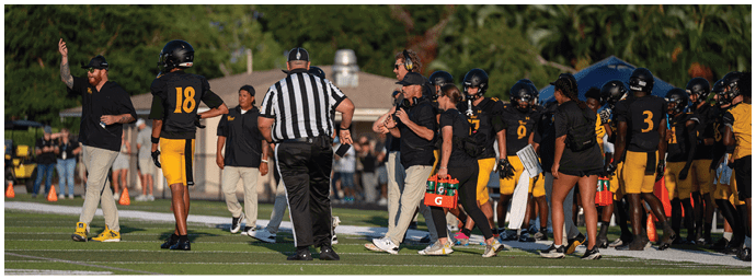 People in sports uniforms and coaches are gathered on the sidelines of a football field during a game or practice. Descripci n generada con IA