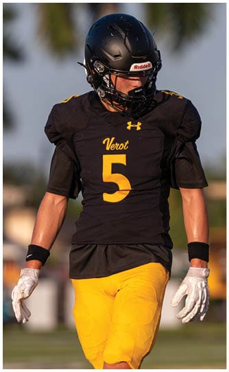 A person wearing a black football helmet and jersey with the number five and yellow pants is standing outdoors with a blurred background. Descripci n generada con IA