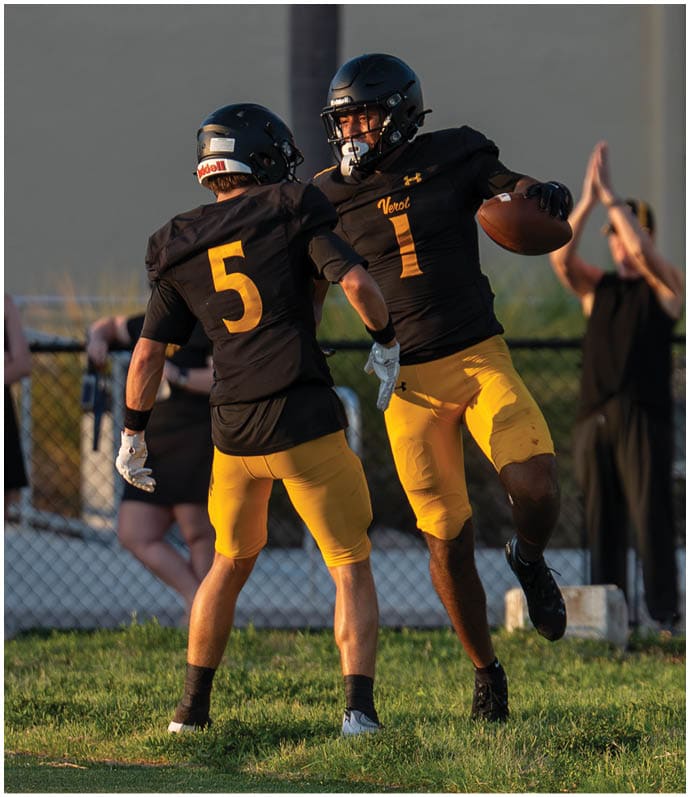 Two people in football uniforms with helmets are celebrating on a field, with one holding a football and the other raising an arm. Descripci n generada con IA