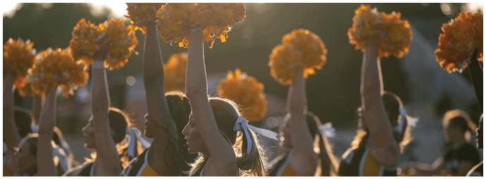 People in uniform with ponytails are raising orange pom-poms in the air during an outdoor event at sunset. Descripci n generada con IA