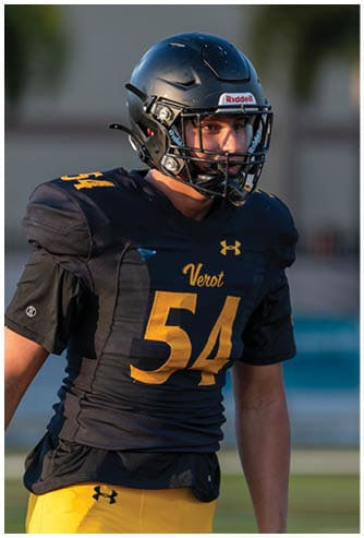 A person wearing a dark football uniform with the number 54 and a helmet is standing outdoors. Descripci n generada con IA