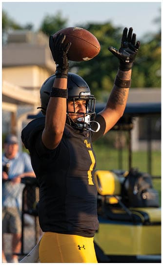 A person wearing a black football uniform and helmet is raising a football above their head on a field with blurred spectators in the background. Descripci n generada con IA