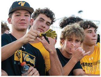 Four young people wearing sports shirts are posing closely together outdoors, with one holding a small foam hand and another making a hand gesture. Descripci n generada con IA