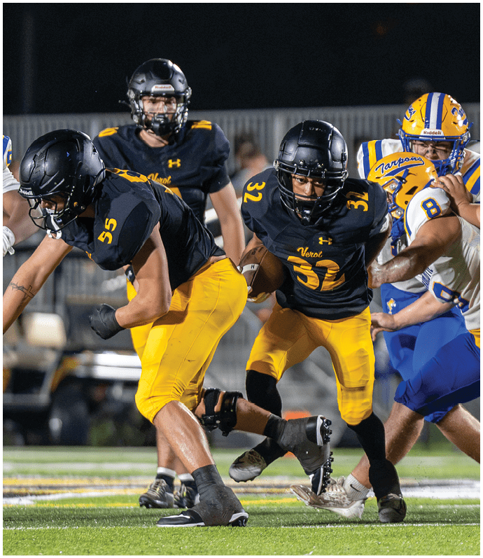 People in football uniforms are actively engaged in a game on a field at dusk, with one player holding the ball and others preparing to block or tackle. Descripci n generada con IA
