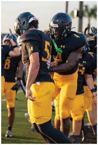 Two people wearing black and yellow football uniforms and helmets are engaged in an intense moment on the field, with one person appearing to shout or encourage the other. Descripci n generada con IA