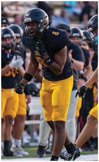 A person wearing a black sports jersey and yellow pants is standing on a football field with clenched fists while other similarly dressed people stand in the background. Descripci n generada con IA