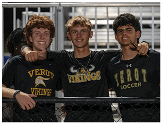 Three adults wearing black shirts with “VEROT VIKINGS" and "VEROT SOCCER" printed on them are smiling and posing together behind a fence at a sports venue. Descripci n generada con IA