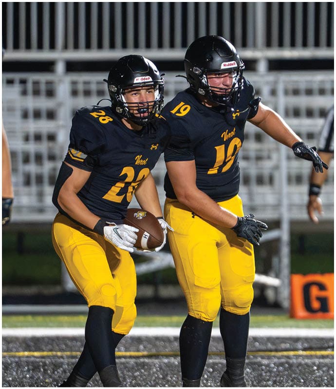 Two people wearing black helmets and black and yellow football uniforms are on a field, with one holding a football and the other appearing to prepare for a play. Descripci n generada con IA