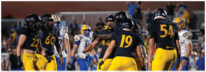 A group of people wearing black helmets and yellow pants are gathered on a football field with others in blue and yellow uniforms in the background. Descripci n generada con IA