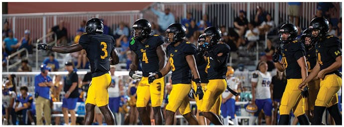 A group of people wearing black helmets and black and yellow sports uniforms are gathered on a field with spectators in the background. Descripci n generada con IA