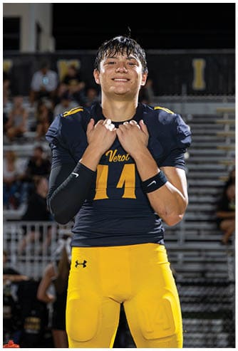 A person wearing a dark blue and yellow football uniform with the number 14 is standing on a field at night, smiling and holding the collar of their jersey. Descripci n generada con IA