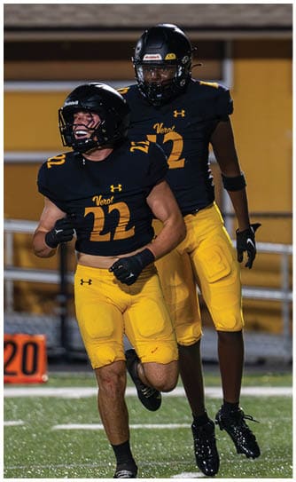 Two people wearing black and yellow football uniforms and helmets are on a field, with one person in the foreground and the other slightly behind. Descripci n generada con IA