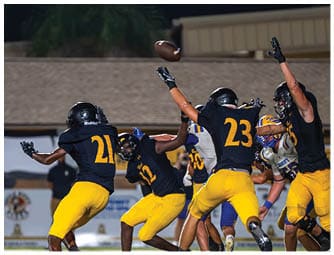 Several people wearing black helmets and yellow pants are actively reaching for a football in mid-air during a nighttime outdoor game. Descripci n generada con IA