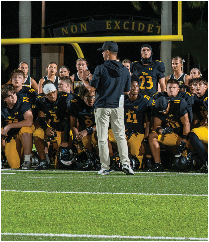 A group of people wearing matching sports uniforms are gathered on a football field at night, listening to a person standing and speaking to them. Descripci n generada con IA