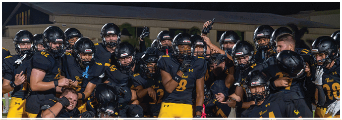 A group of people wearing black helmets and yellow pants are gathered closely together on a sports field at night, with one person in the center raising a hand. Descripci n generada con IA
