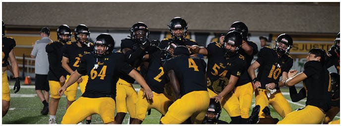 A group of people wearing black and yellow football uniforms are gathered on a field at night, some standing and some sitting, appearing to be in a team huddle or discussion. Descripci n generada con IA