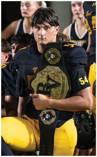A person wearing a black football uniform holds a large championship belt with intricate gold detailing and the letters RV while standing among other people in similar uniforms. Descripci n generada con IA