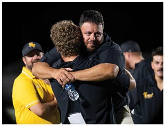 Two adults wearing black shirts embrace tightly on a sports field at night while other people in sports attire watch nearby. Descripci n generada con IA