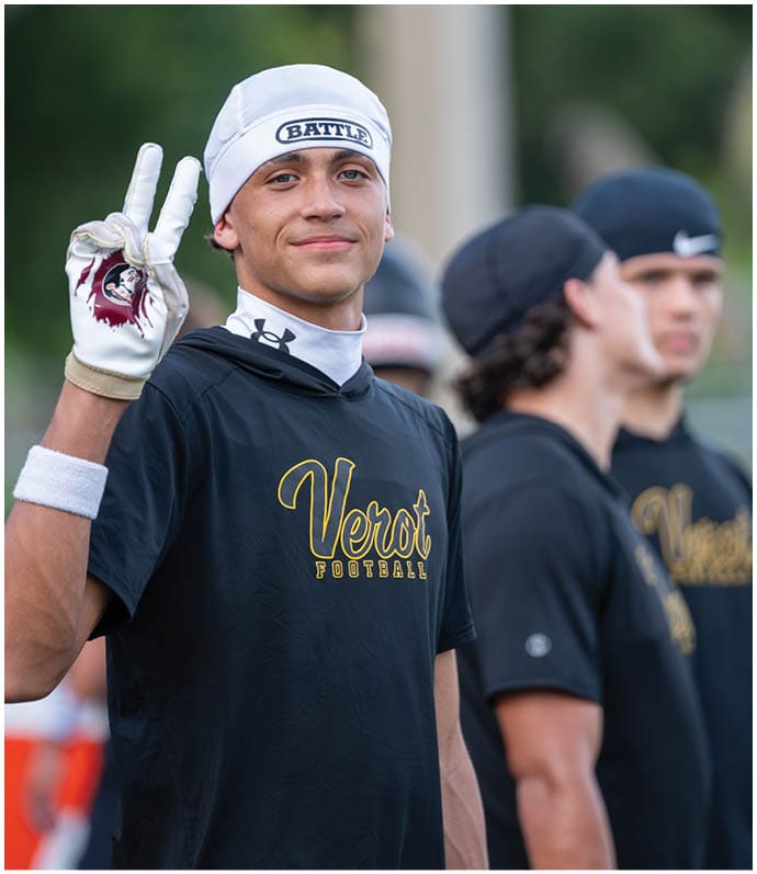 A person wearing a white headband and black shirt with “Verot Football" printed on it is smiling and holding up a peace sign while standing outdoors with other people in similar attire blurred in the background. Descripci n generada con IA