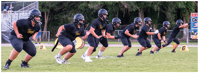 Several people wearing black athletic clothing and helmets are lined up in a crouched position on a grassy field, appearing ready to start a race or drill. Descripci n generada con IA