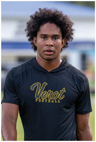 A person with curly hair wearing a black hoodie is looking forward with a neutral expression against a blurred outdoor background. Descripci n generada con IA