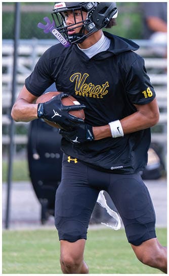 A person wearing a black football helmet and uniform is holding a football and appears to be preparing to throw it on a grassy field with bleachers in the background. Descripci n generada con IA
