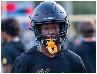 A person wearing a black football helmet and black sports jersey with yellow lettering is looking directly at the camera while holding an orange mouthguard in their mouth. Descripci n generada con IA