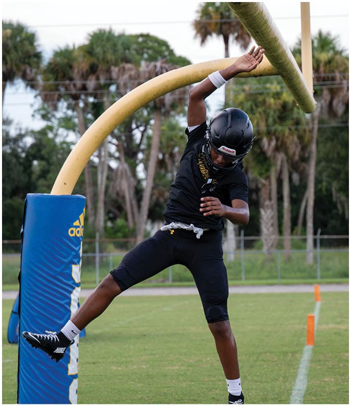 A person wearing a black helmet and sportswear is jumping and reaching up to touch a yellow football goalpost on a grassy field with palm trees in the background. Descripci n generada con IA