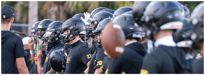 A group of people wearing black football helmets and uniforms are gathered outdoors with a coach giving instructions. Descripci n generada con IA
