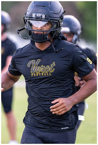 A person wearing a black football helmet and black Verot Football uniform is running on a grassy field with other players in the background. Descripci n generada con IA