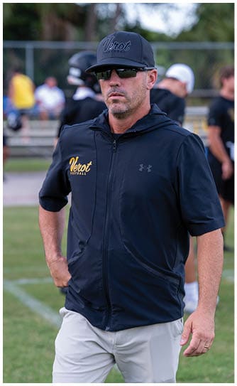 A person wearing a black cap and sunglasses with a black zip-up jacket that has “Verot Football" written on it stands outdoors with blurred people and trees in the background. Descripci n generada con IA