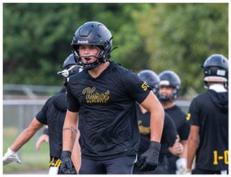 A person wearing a black football helmet and black shirt with “Verot" written on it is on a football field with other similarly dressed people in the background. Descripci n generada con IA