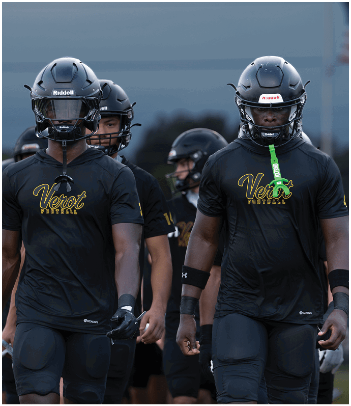 People wearing black football helmets and black shirts with “Verot Football" written on them are walking outdoors with a football in midair above them. Descripci n generada con IA