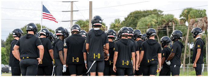 A group of people wearing black sports uniforms and helmets are gathered closely together on a grassy field, likely preparing for a team activity or game. Descripci n generada con IA
