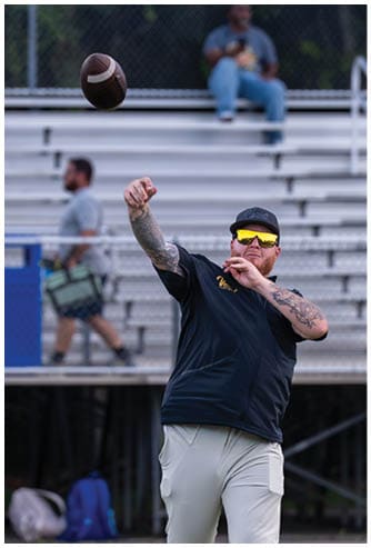 A person wearing sunglasses, a cap, a dark shirt, and light pants is throwing a football on a field with empty bleachers and two other people in the background. Descripci n generada con IA