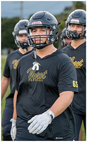 A person wearing a gray football helmet with a black faceguard is focused ahead with another person in a similar helmet blurred in the background. Descripci n generada con IA