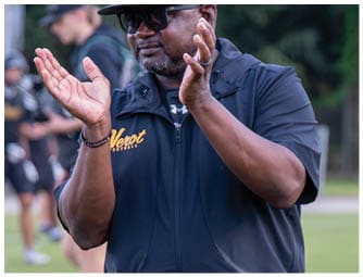 A person wearing a dark cap, sunglasses, and a dark zip-up jacket is clapping on a sports field with other people and sports equipment blurred in the background. Descripci n generada con IA