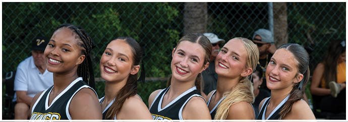 Five adults with braided and tied-back hair are smiling closely together outdoors in front of a chain-link fence with trees in the background. Descripci n generada con IA