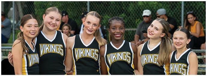 Six smiling people wearing matching black and yellow Vikings cheerleading uniforms stand closely together outdoors with a chain-link fence and seated people in the background. Descripci n generada con IA