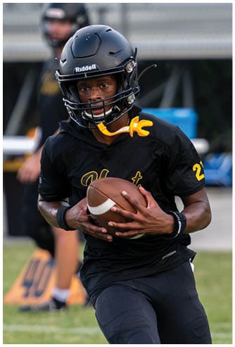A person wearing a black football helmet and jersey is holding a football and appears to be preparing to throw it during a game or practice. Descripci n generada con IA