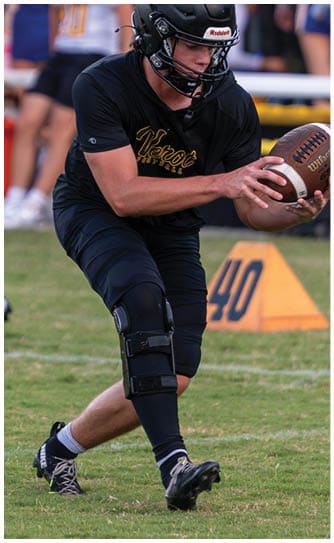 A person wearing a black football helmet and black athletic clothing is crouched on a grassy field near a marker with the number 40, with blurred people and sports equipment in the background. Descripci n generada con IA