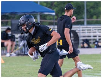An adult wearing a black football uniform and helmet is running with a football while another adult in black athletic clothing walks in the background on a grassy field. Descripci n generada con IA