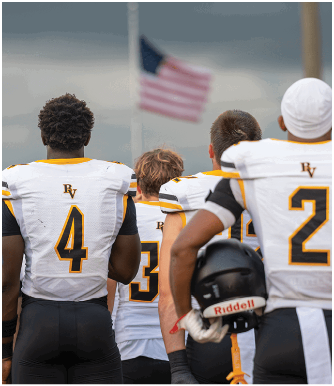 Three people wearing white and yellow sports jerseys stand with their backs to the camera facing a blurred American flag on a flagpole against a cloudy sky. Descripci n generada con IA