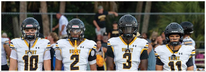 Four people wearing football helmets and white jerseys with black and yellow accents stand side by side on a field with a chain-link fence and trees in the background. Descripci n generada con IA