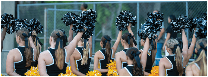 A group of people in matching black and white cheerleading uniforms hold black and yellow pom-poms, standing outdoors near a chain-link fence. Descripci n generada con IA