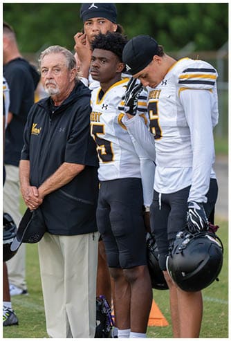 A senior person in a black jacket stands next to two people in white and yellow football uniforms, one looking forward and the other looking down while adjusting a glove. Descripci n generada con IA