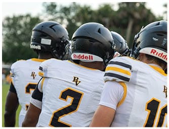 Four people wearing white and yellow football uniforms and black helmets stand closely together on a grassy field with trees in the background. Descripci n generada con IA