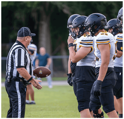 A referee in a striped shirt is holding a football and speaking to three football players wearing helmets and uniforms on a field. Descripci n generada con IA